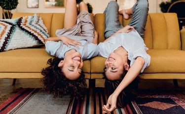 Zwei Frauen liegen kopfüber auf einem Sofa und lachen.